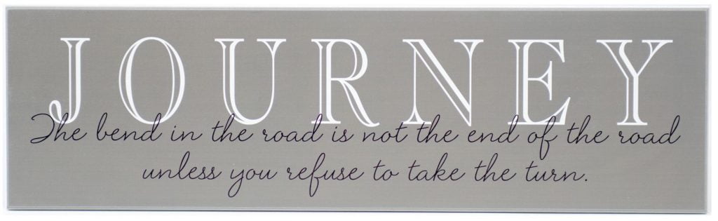 Charcoal wooden sign with saying, "Journey-The bend in the road is not the end of the road unless you refuse to take the turn" in black through the middle of the sign.